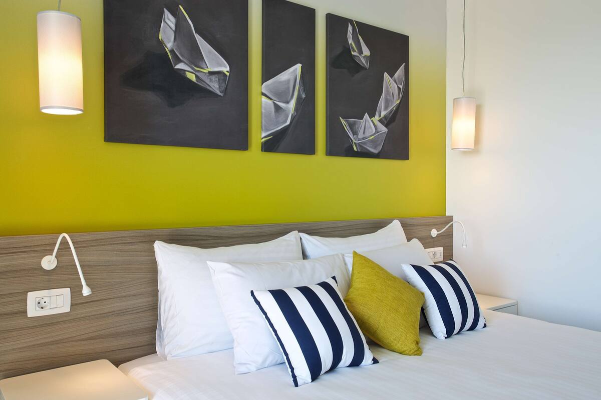 Quarto de hotel com cama, almofadas, mesa de cabeceira, tomada e três obras de arte na parede.