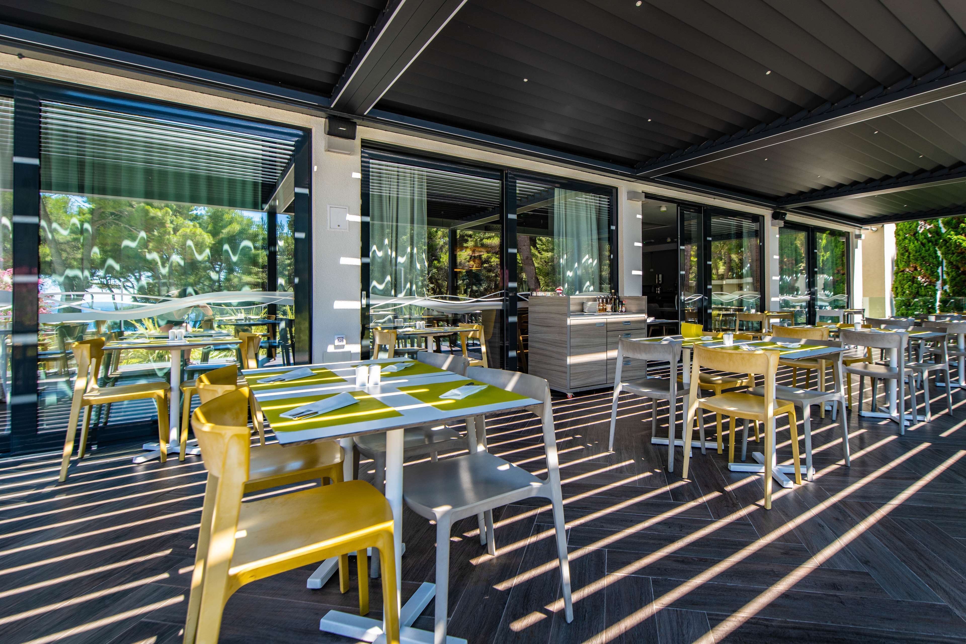 An outdoor dining area with modern furniture and large windows offering views of greenery.