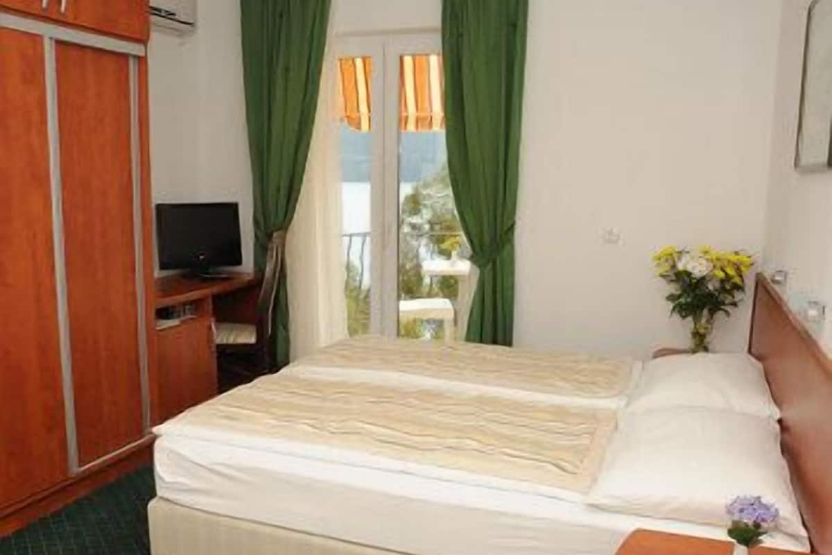 A hotel room with a bed, a desk with a television, a window with curtains, and a nightstand with a vase of flowers.