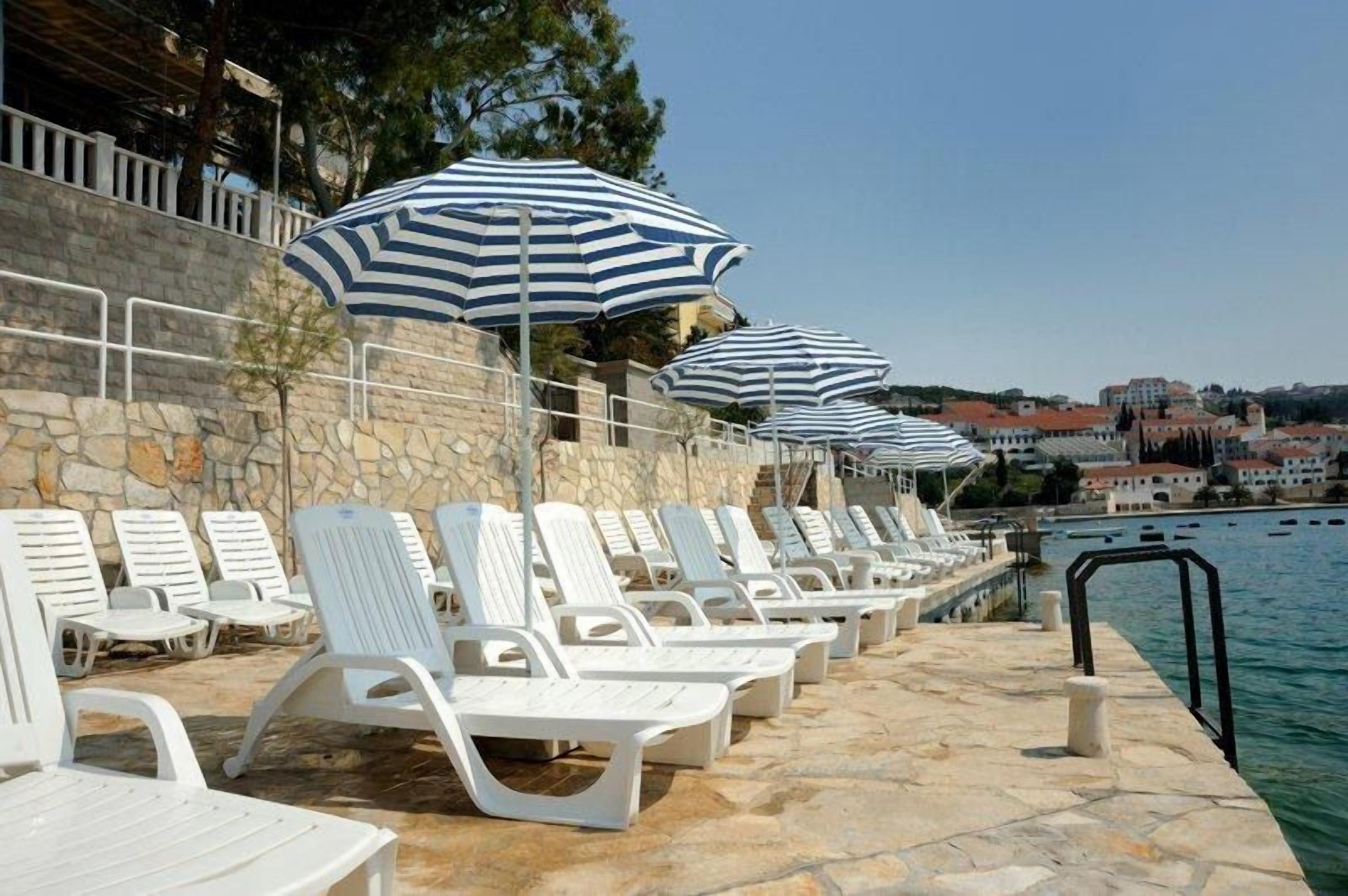 A row of white lounge chairs and striped umbrellas by a stone wall and a body of water.