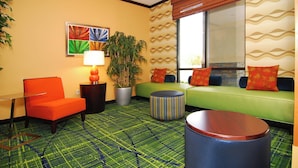 Habitación de hotel con un sofá verde, un sillón naranja y un reposapiés a rayas. Hay una planta en maceta y una lámpara sobre una mesita de noche.