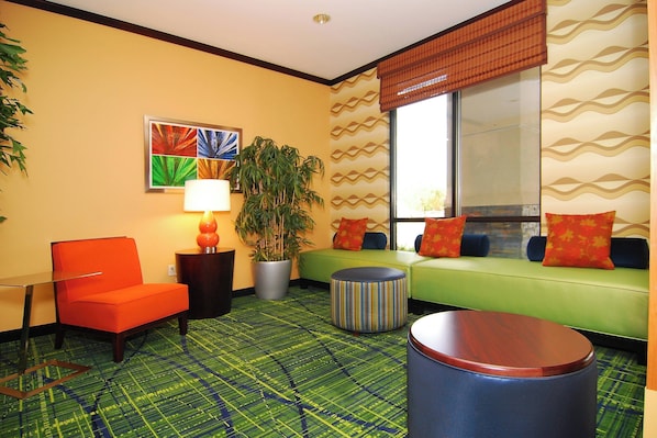A hotel room with a green sofa, orange armchair, and a striped ottoman. There is a potted plant and a lamp on a side table.