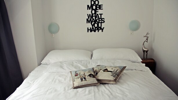 Un lit bien fait, avec du linge de lit blanc et deux coussins décoratifs, un mur orné d’une citation inspirante et une petite table sur laquelle est posé un objet décoratif.