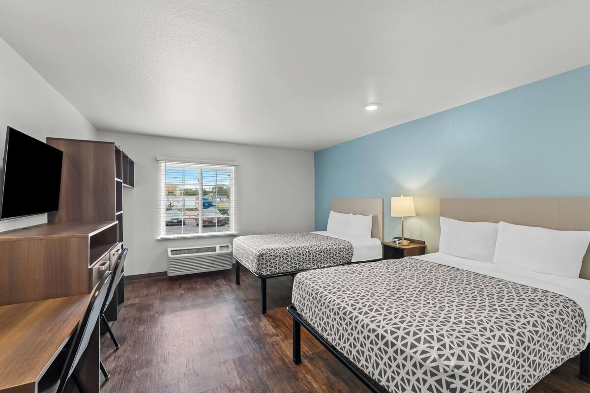 A hotel room with a large bed, a smaller bed, a desk, a TV, and a window with blinds.