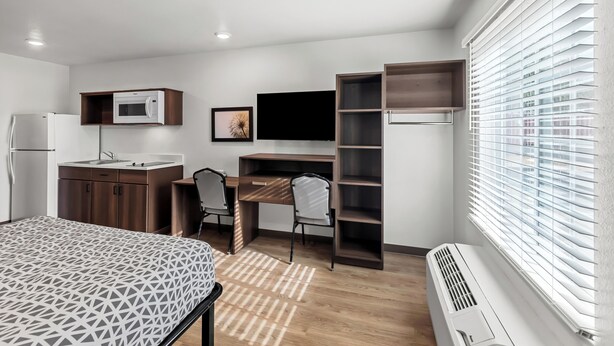 Une chambre moderne comprenant un lit, un bureau, une chaise, une télévision, une armoire et une fenêtre avec des stores.
