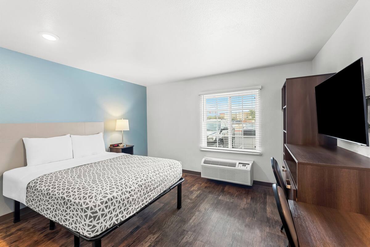 A hotel room with a bed, a desk, a chair, a TV, a window with blinds, and an air conditioning unit.