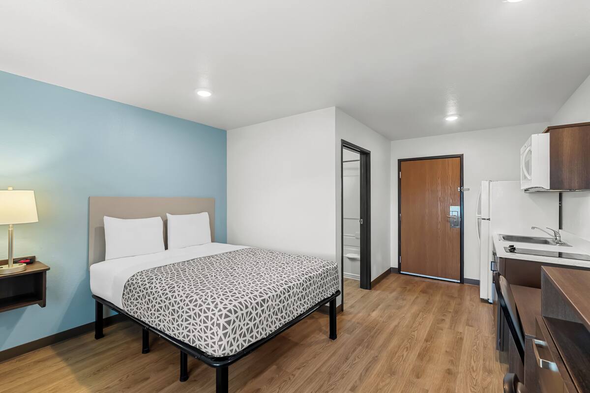 A hotel room with a bed, bedside table, lamp, and a kitchenette area with a microwave and sink.