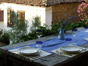 An outdoor dining table set with plates, cutlery, and glasses, surrounded by greenery and a rustic building.