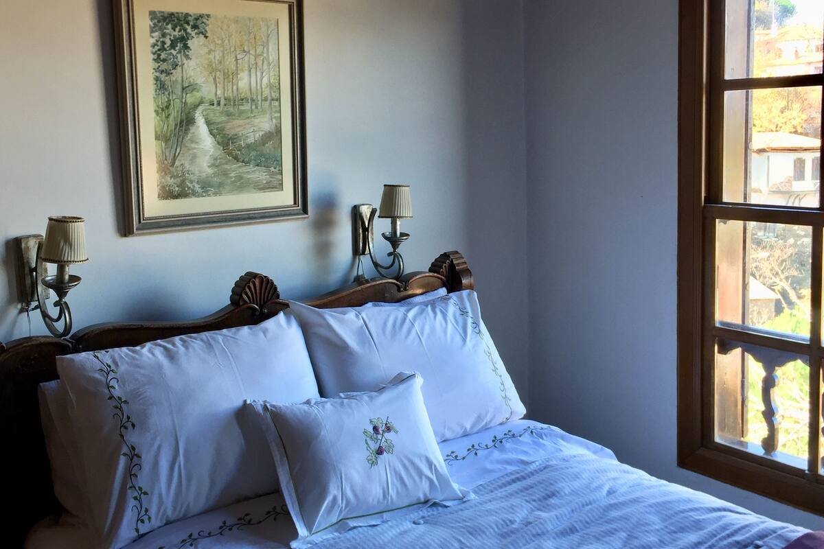 A bedroom with a wooden headboard, white bedding, a framed painting of a forest, two wall-mounted lamps, and a window with a view of trees.
