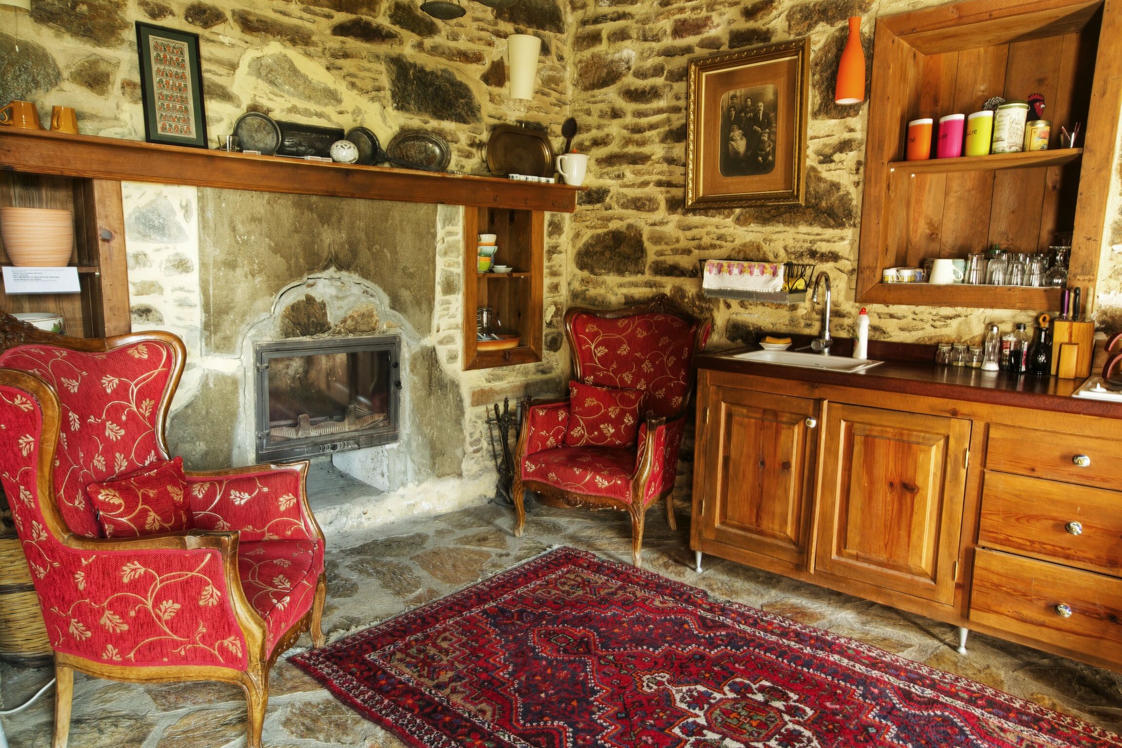 A stone-walled room with a fireplace, red upholstered chairs, a wooden cabinet, and a red patterned rug.