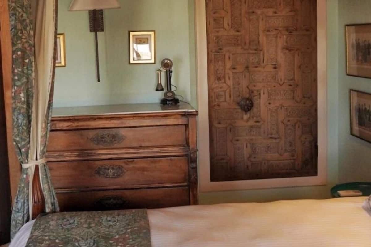 A bedroom with a wooden dresser, a bed with a floral bedspread, a wooden door with intricate patterns, a wall-mounted lamp, and a framed picture.