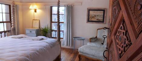 A bedroom with a large bed, a chair, a small table, a lamp, and a painting on the wall.