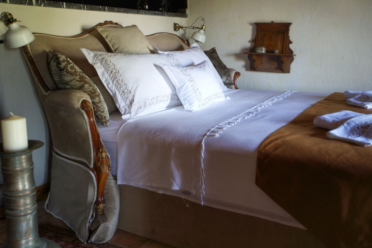 A neatly made bed with white linens and a brown throw blanket, a bedside lamp, and a small wooden shelf with decorative items.