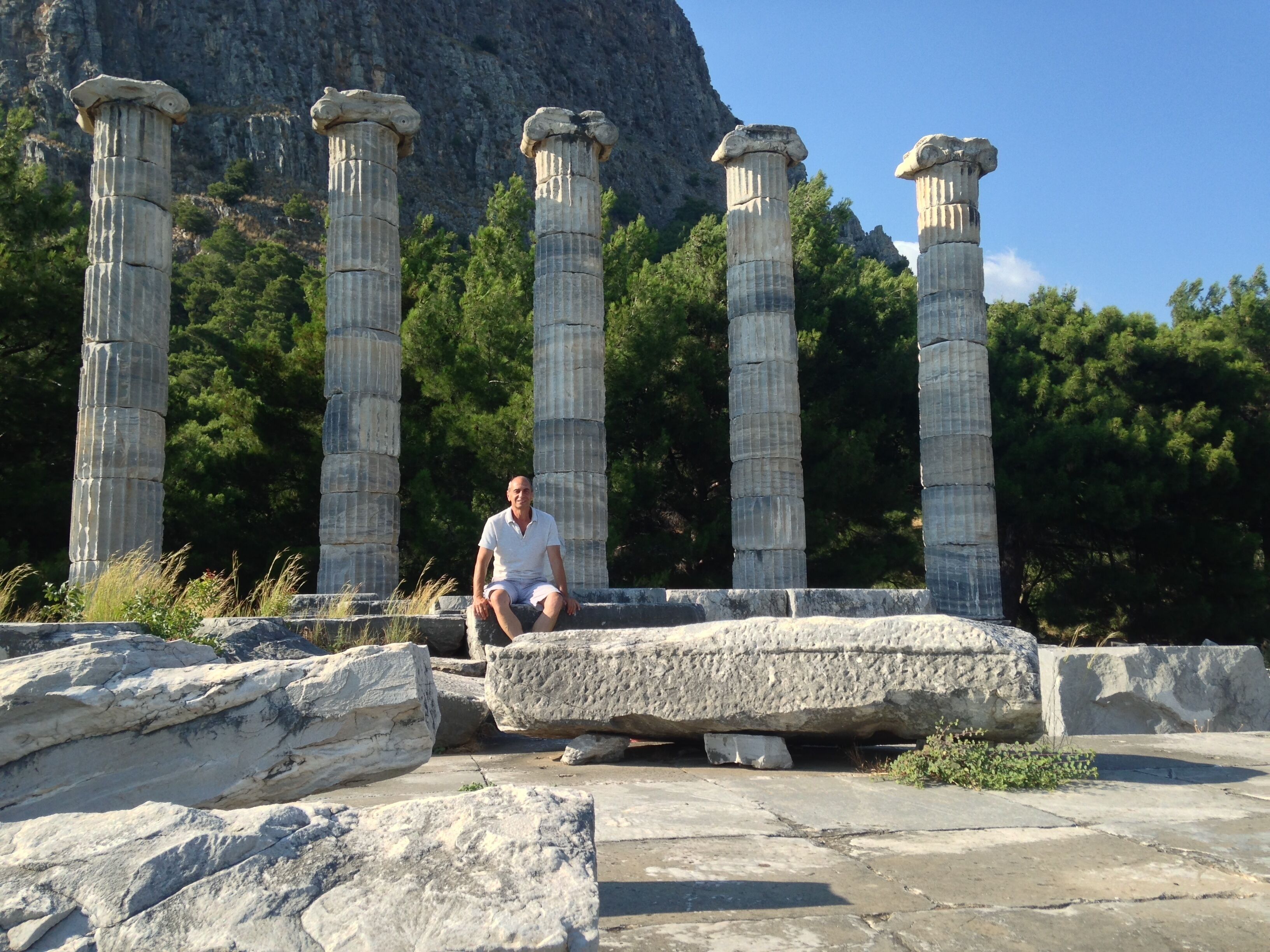 A man sitting on a stone platform with ancient columns and a mountainous backdrop.