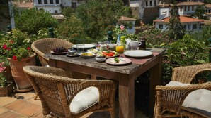 A wooden table set for a meal with wicker chairs, surrounded by potted plants and overlooking a hillside of traditional buildings. - Terrace Houses Sirince - Fig, Olive Clockmakers and Grapevine (Selçuk)