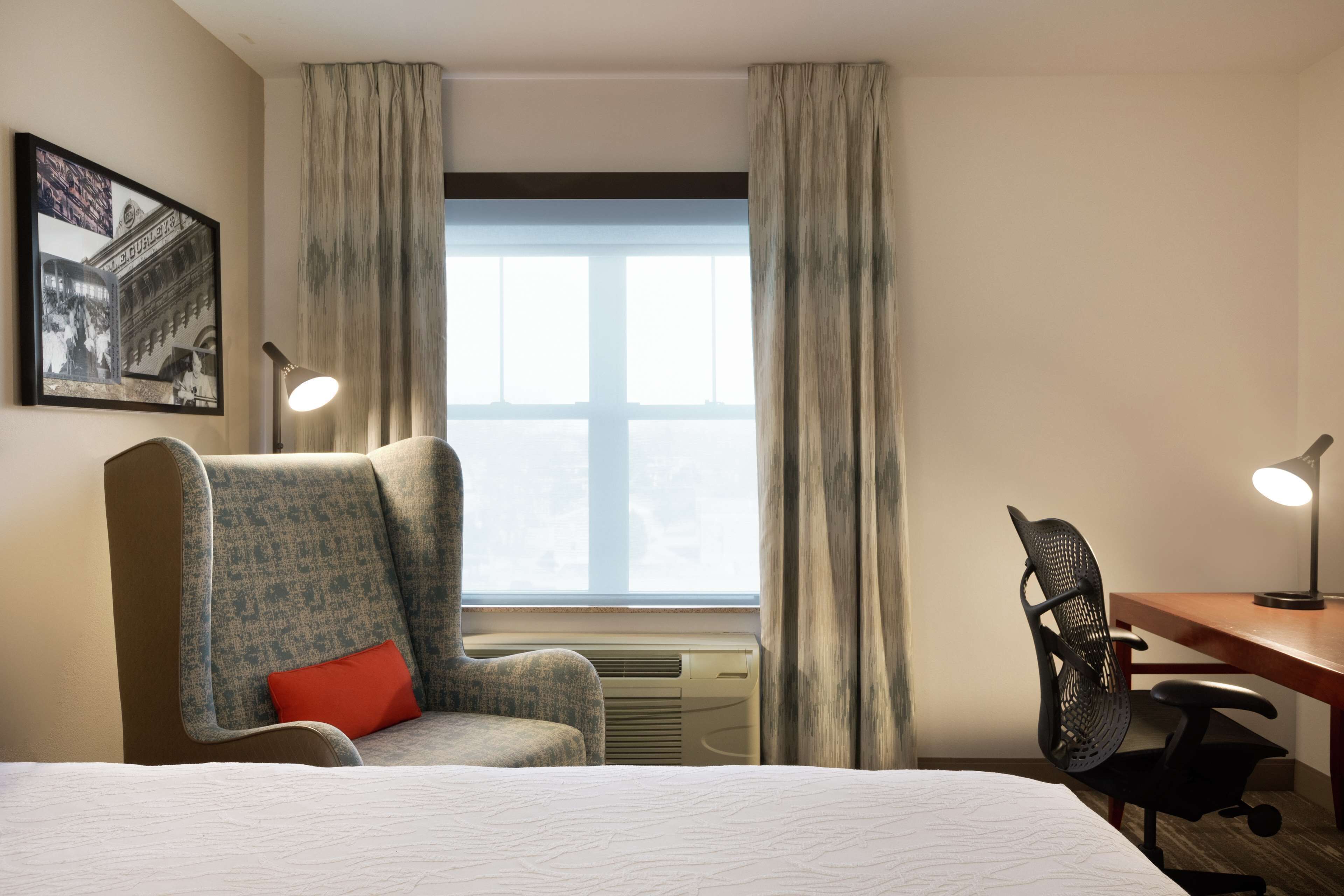 Una habitación de hotel con cama, escritorio, dos sillas, ventana con cortinas, aire acondicionado y un cuadro en la pared.