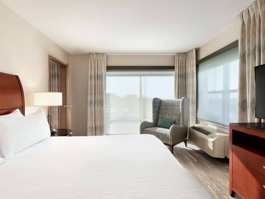 A hotel room with a large bed, a chair, a TV, and a window with curtains.