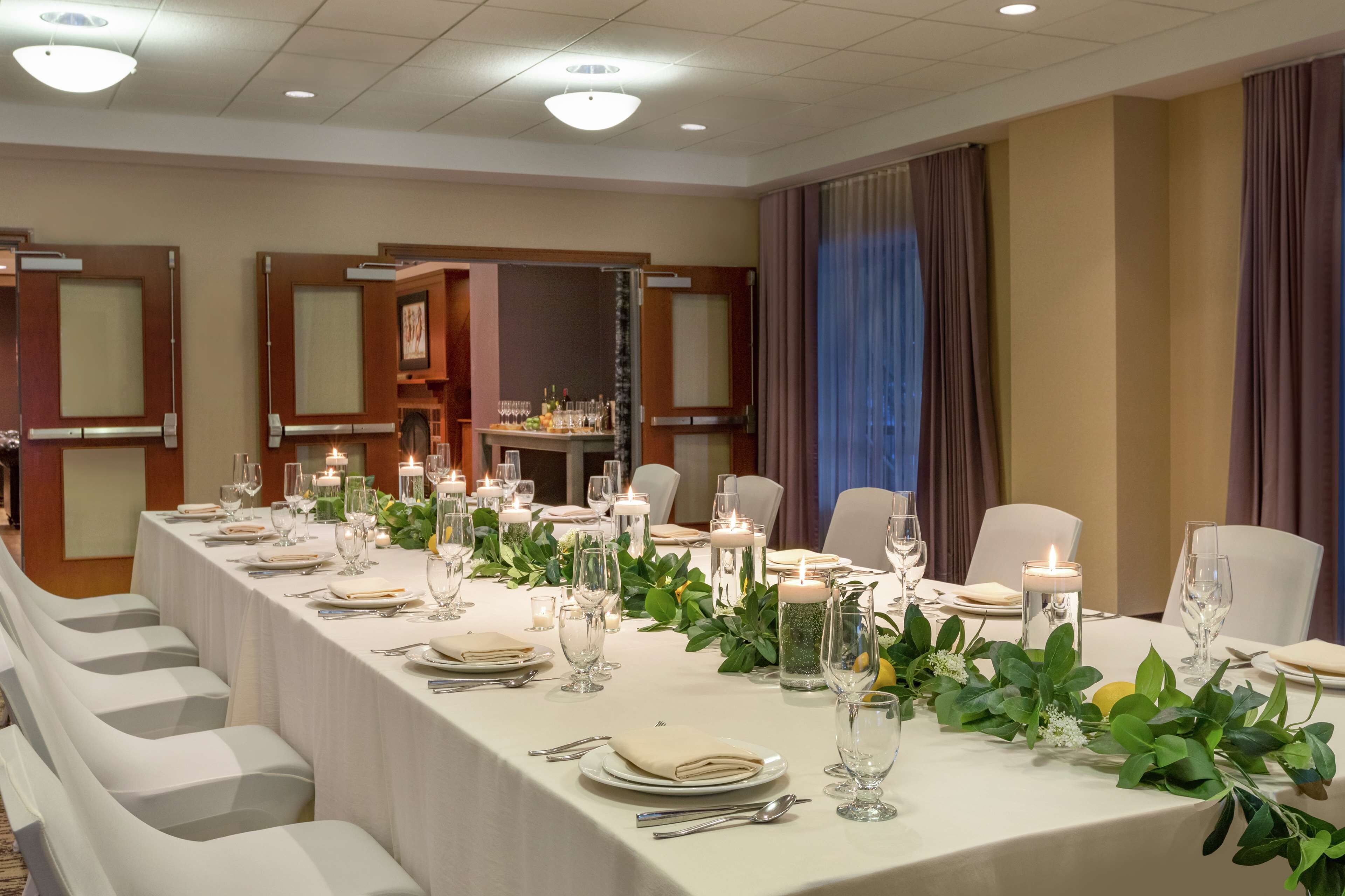Una larga mesa de banquete dispuesta con manteles blancos, velas y follaje, rodeada de sillas blancas en una habitación bien iluminada con puertas de madera y un gran ventanal con cortinas.