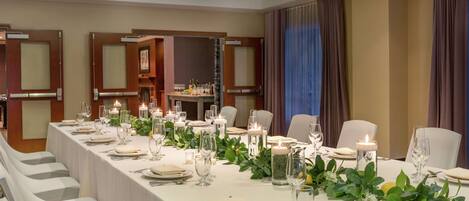 Una larga mesa de banquete dispuesta con manteles blancos, velas y follaje, rodeada de sillas blancas en una habitación bien iluminada con puertas de madera y un gran ventanal con cortinas.