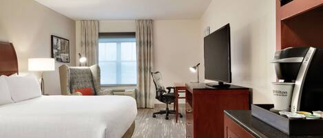 Habitación de hotel con cama, escritorio, silla, televisor, minibar y ventana con cortinas.