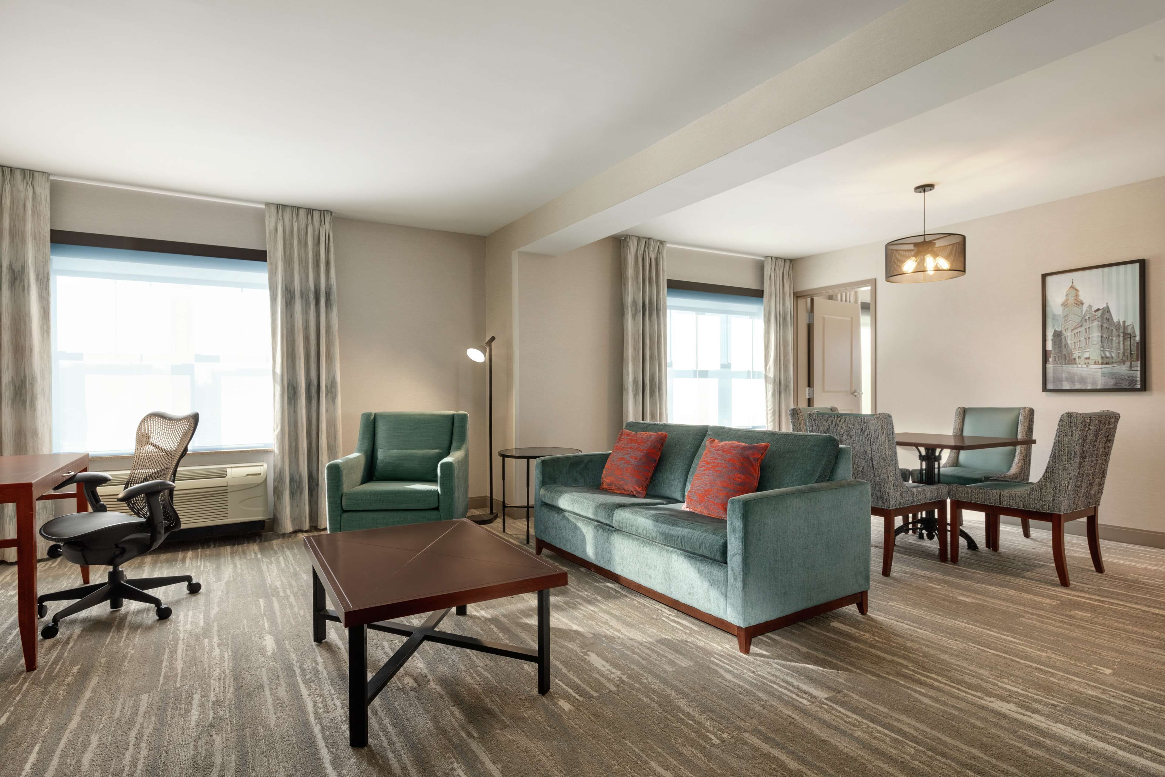 A hotel room with a green sofa, a wooden coffee table, a dining table with chairs, a standing lamp, and a large window with curtains.