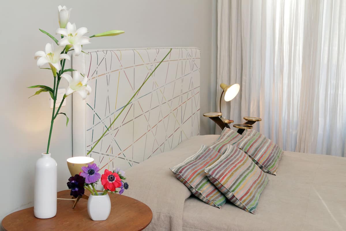 A hotel room with a bed, striped pillows, a bedside table with a vase of flowers, and a wall with a geometric pattern.