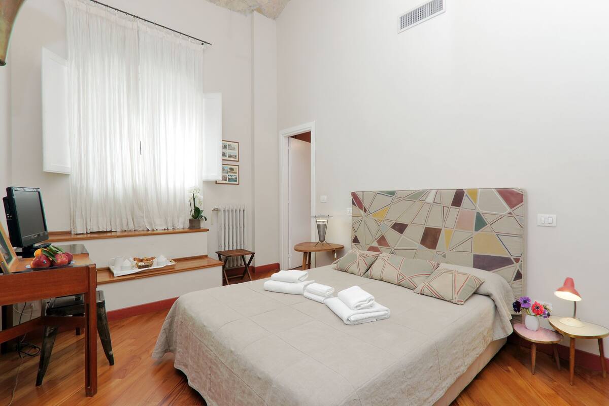 A hotel room with a bed, a desk, a chair, a small table, a lamp, and a window with curtains.
