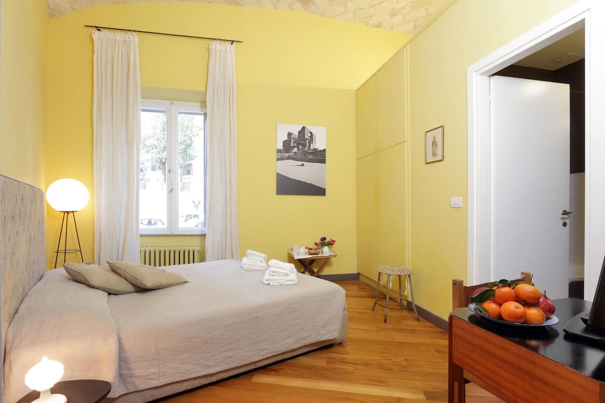 A hotel room with a bed, a small table with a fruit bowl, a chair, a lamp, and a picture on the wall.