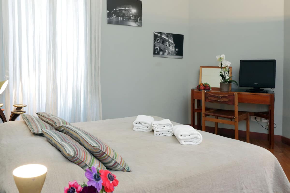 A neatly made bed with pillows and folded towels, a wooden desk with a computer, and a potted plant.