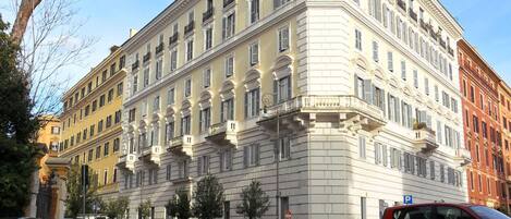 A large, multi-story building with classical architecture, featuring a central courtyard and a parking area in front.