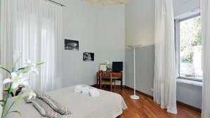 Habitación de hotel con cama, escritorio con computadora, lámpara de pie y ventana con cortinas.
