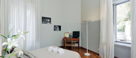 Habitación de hotel con cama, escritorio con computadora, lámpara de pie y ventana con cortinas.