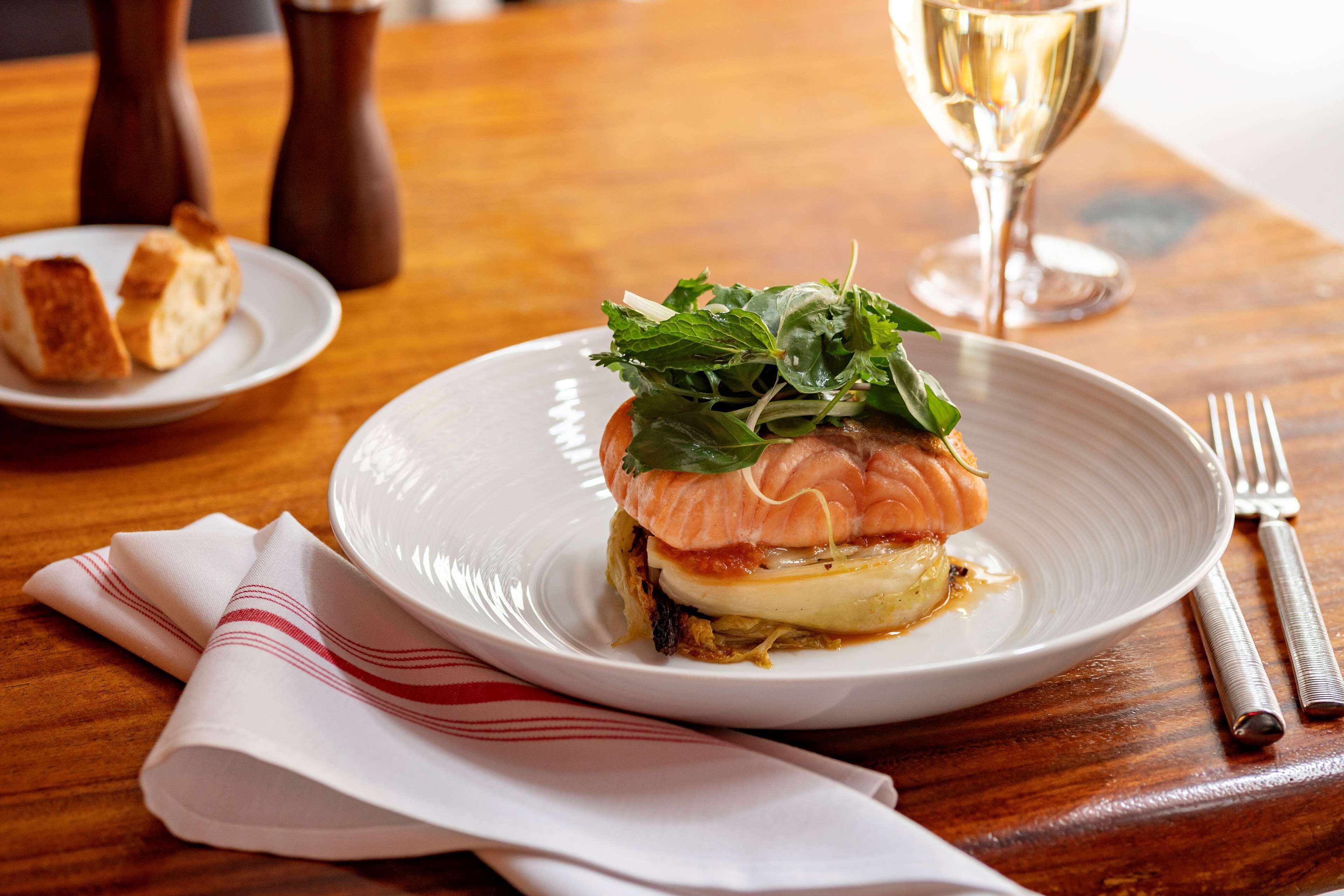 A plate with a sandwich, a glass of white wine, and a folded napkin on a wooden table.