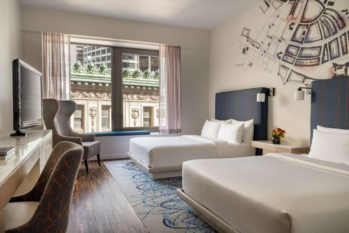 Habitación de hotel con cama, escritorio con televisor, silla y ventana con vista a edificios.