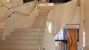 Un escalier moderne avec des rampes en verre et des marches en bois, menant à un hall d’entrée doté d’un mur en bois et d’un espace salon.