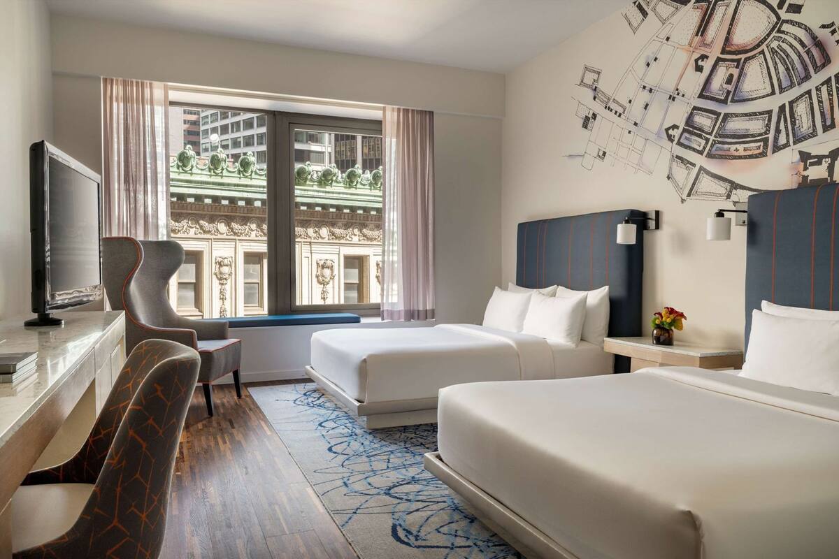 Habitación de hotel con dos camas, un escritorio con televisor, una silla y un ventanal con vistas a edificios.