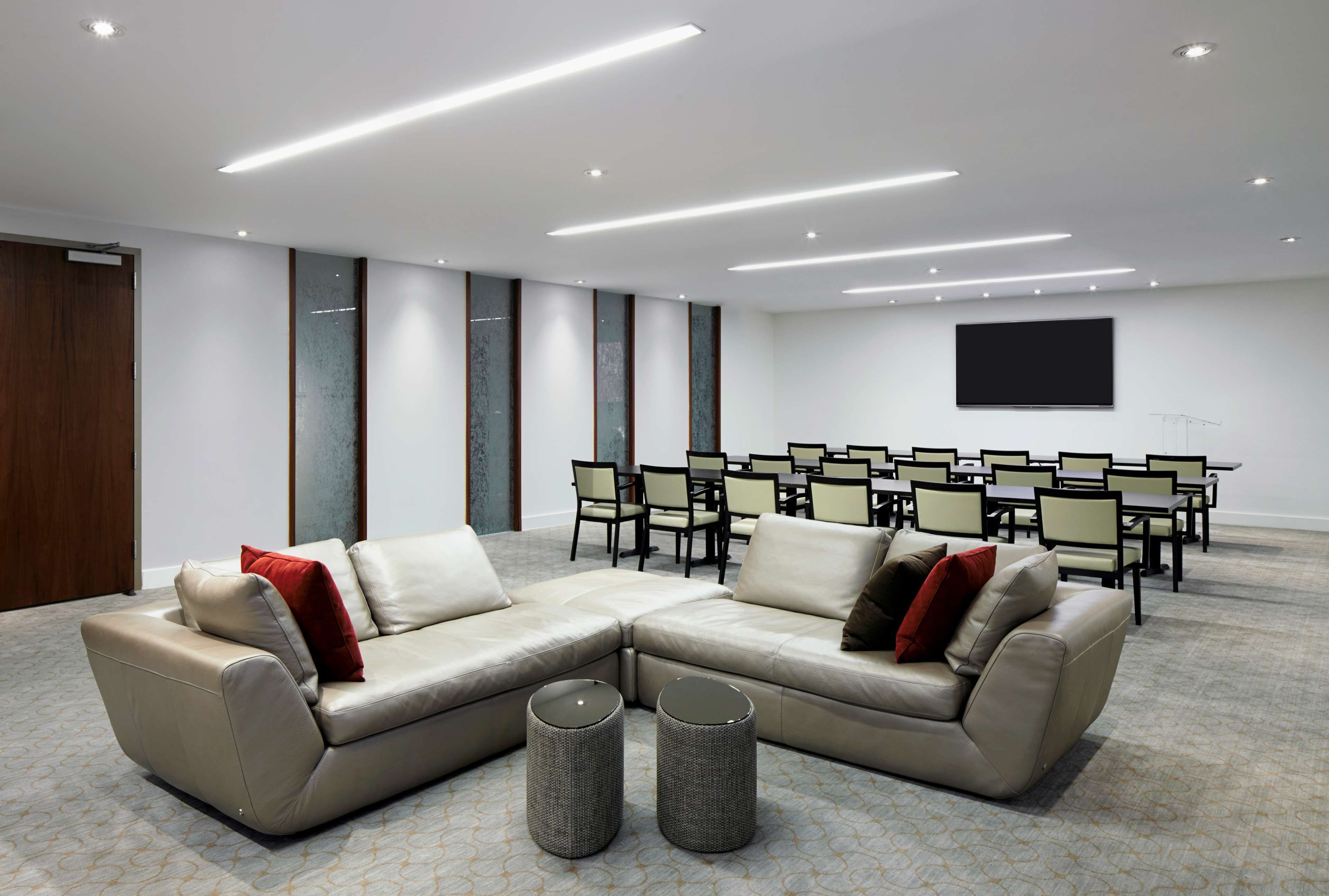 A conference room with a large L-shaped sofa, two round side tables, and a row of chairs facing a flat-screen TV.