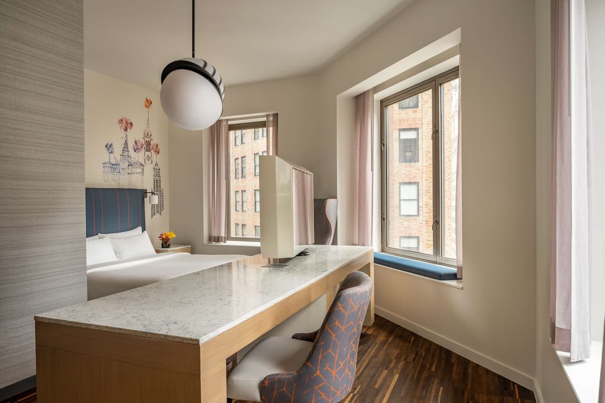 Habitación de hotel con cama, escritorio, silla, ventana con vista a edificios y pared decorada con un mural.