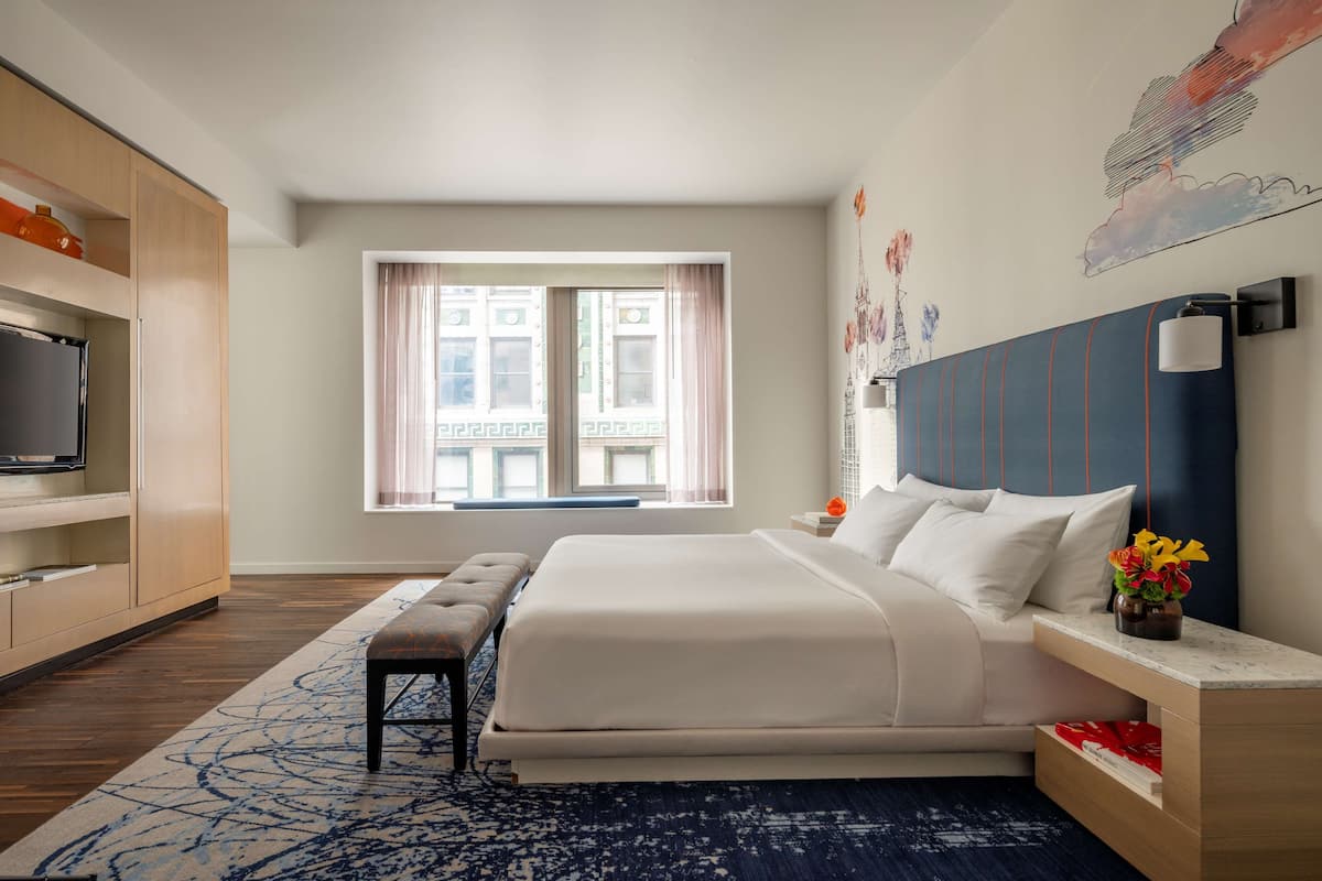 Habitación de hotel moderna con una cama grande, televisión, un banco y vistas de la ciudad a través de la ventana.