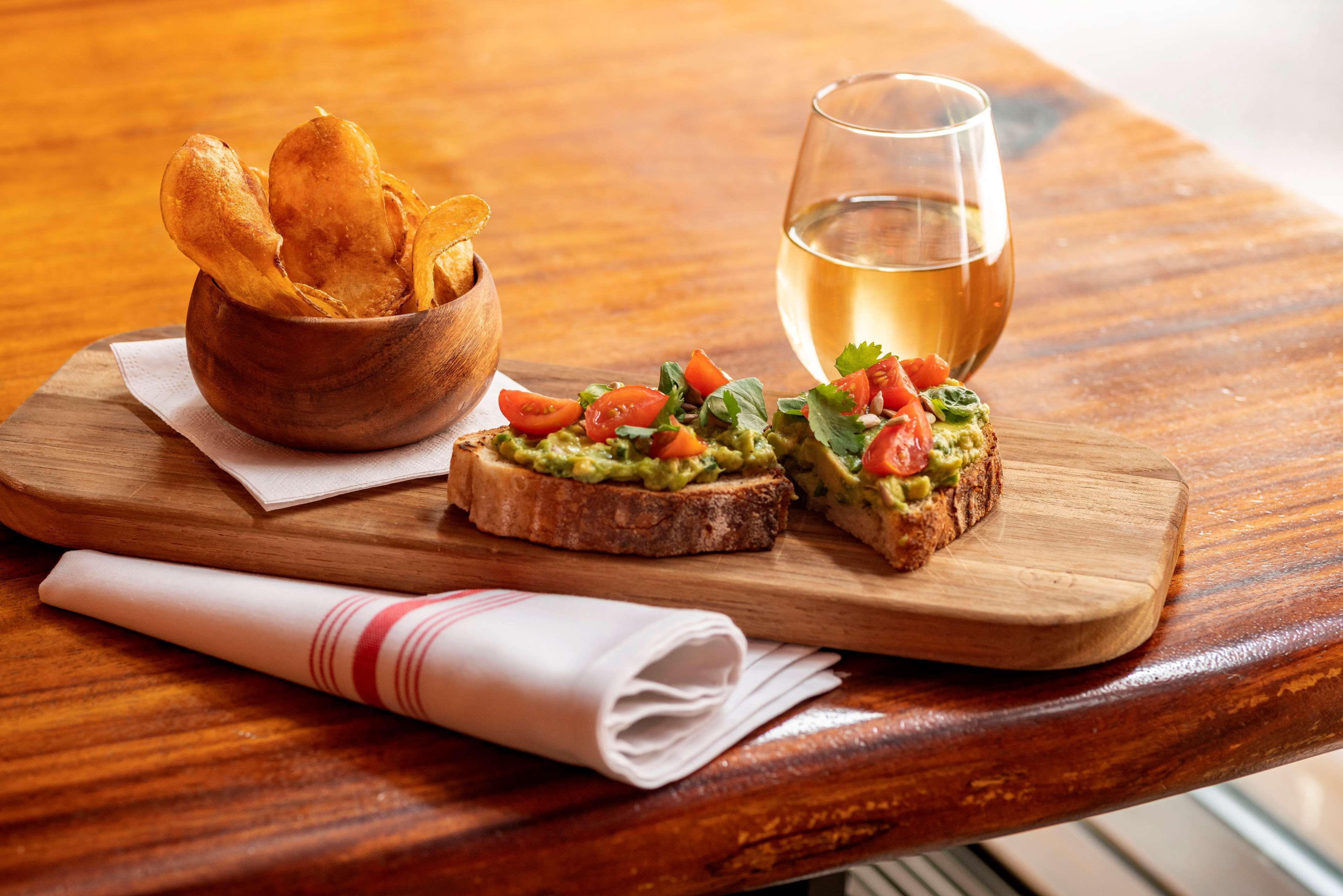 A wooden table with a wooden bowl of chips, two pieces of toast with avocado and tomato, and a glass of white wine.