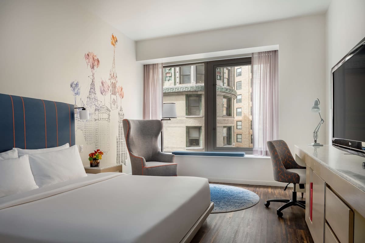 Habitación de hotel con una cama grande, un escritorio con televisor, una silla y ventana con vista a edificios.
