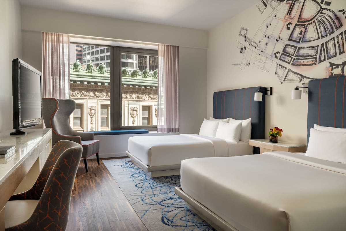 Habitación de hotel con dos camas, un escritorio, una silla y un ventanal con vistas a un edificio.