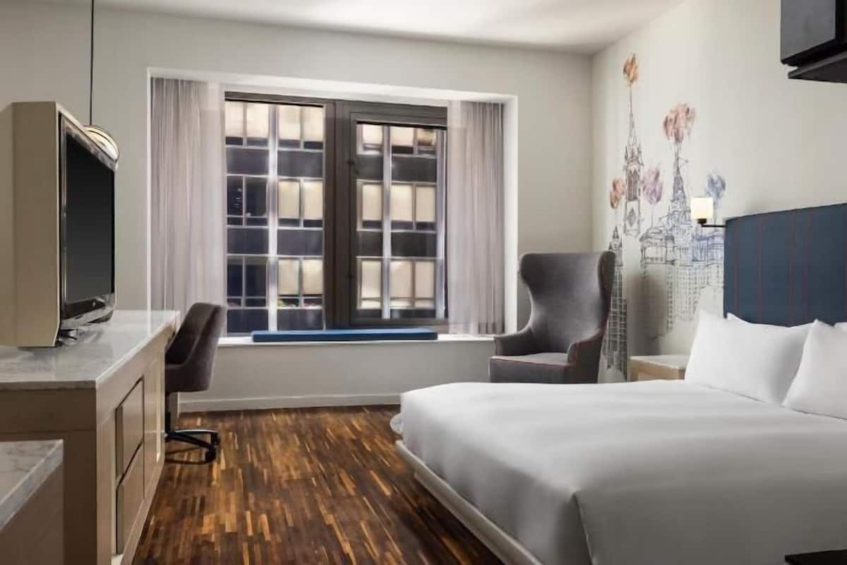 Habitación de hotel con cama, escritorio con televisor, silla, ventana con cortinas y mural de paisaje urbano en la pared.