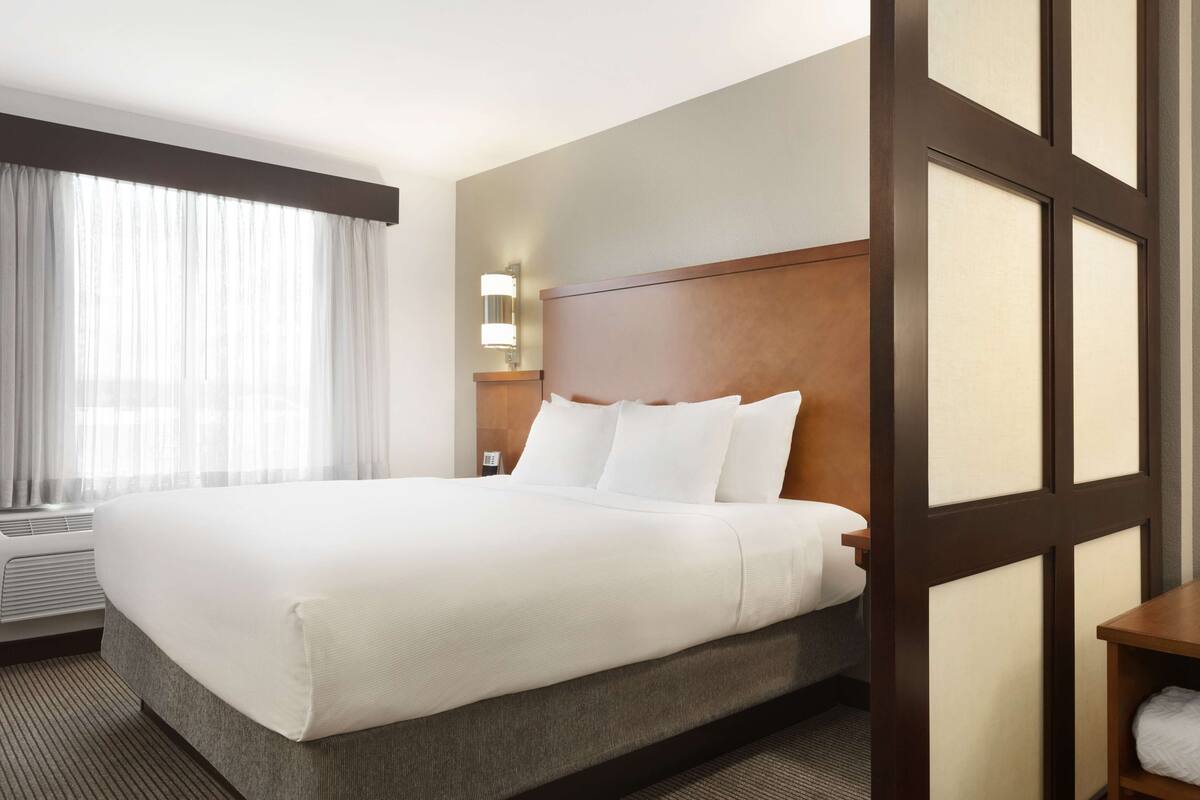A hotel room with a large bed, a nightstand with a lamp, a window with sheer curtains, and a wooden headboard.