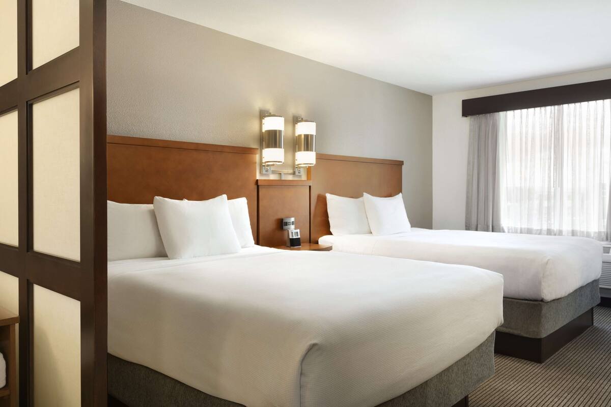 A hotel room with two beds, a wooden headboard, a nightstand, and a window with sheer curtains.