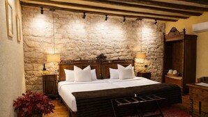A bedroom with a stone wall, wooden bed frame, white bedding, two bedside lamps, a red rug, and a wooden bench.