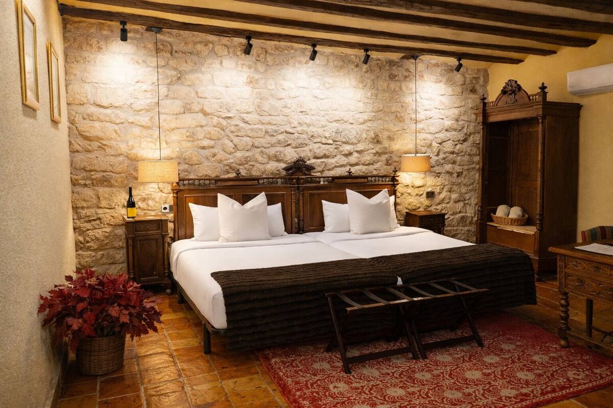 A bedroom with a stone wall, wooden bed frame, white bedding, two bedside lamps, a red rug, and a wooden bench.