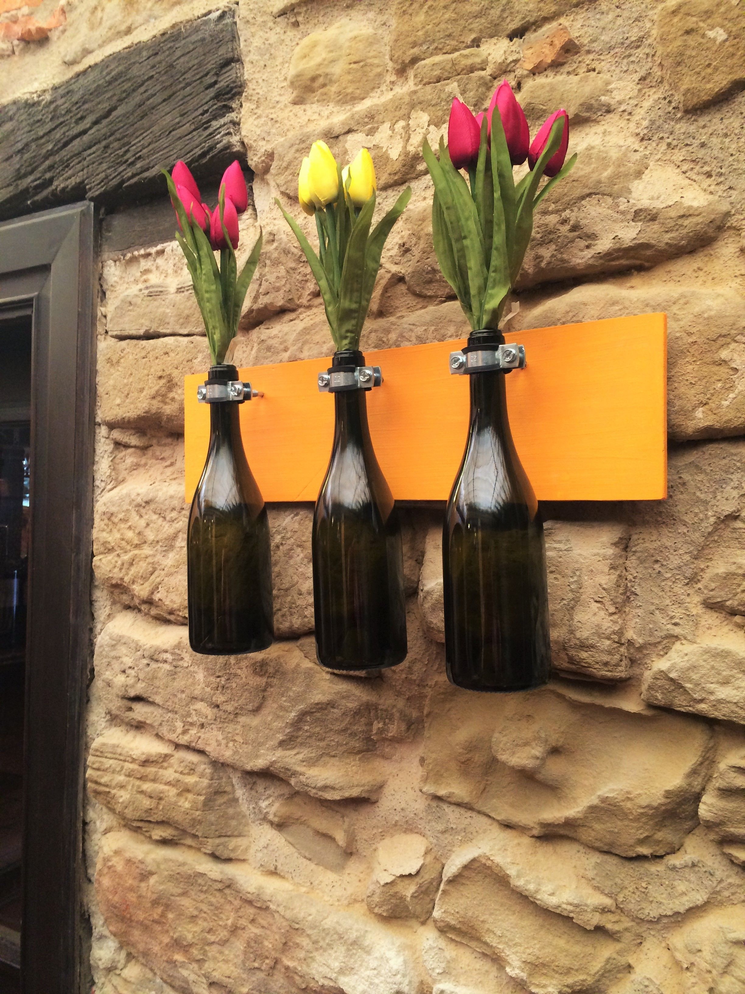 Three glass bottles with flowers against a stone wall.