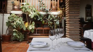 A dining table set with plates, cutlery, and wine glasses, surrounded by potted plants and a brick column.