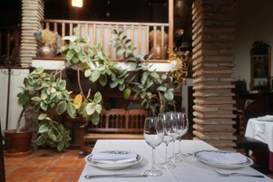 Mesa de jantar posta com pratos, talheres e copos de vinho, cercada por plantas em vasos e um pilar de tijolos.